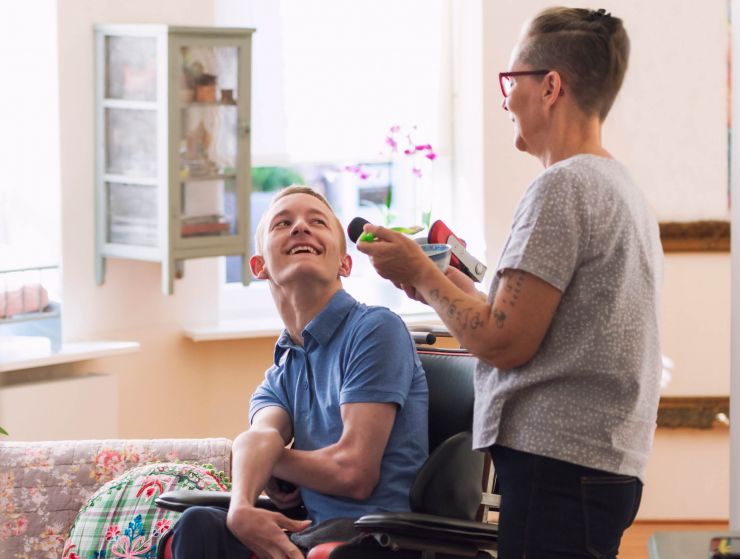Photo of two people in a bright living room smiling at each other. One is a wheelchair user with short hair and a blue shirt. The other has short hair and glasses and is standing.